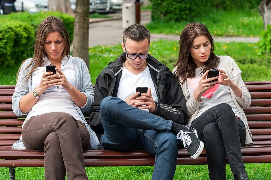 公園のベンチに座って、携帯電話をいじっている3人の人々。