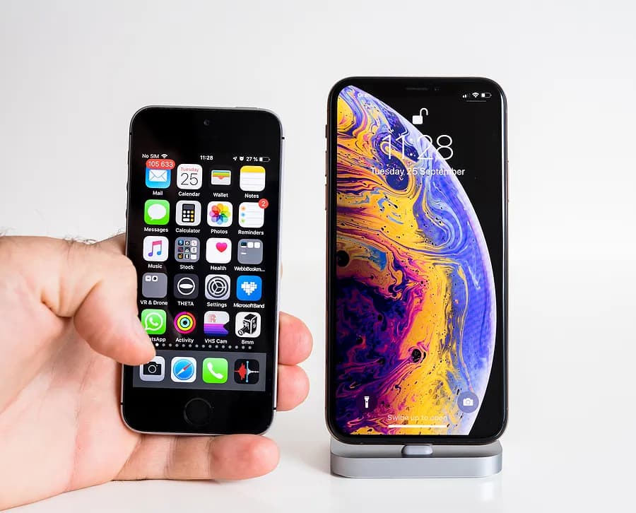 スタンドに置かれた大型の最新型iPhoneの隣に、小型の旧型iPhoneを手に持った写真。これにより、旧型iPhoneと新型モデルのサイズやデザインの違いがわかる。
